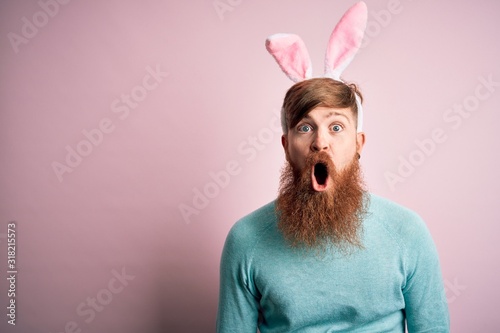 Canvas Print Hipster Irish man with beard wearing easter rabbit ears over isolated pink background afraid and shocked with surprise expression, fear and excited face