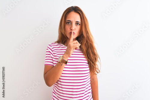 Young redhead woman wearing striped casual t-shirt stading over white isolated background asking to be quiet with finger on lips. Silence and secret concept.