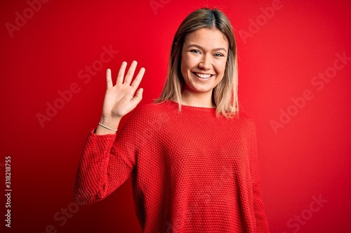 Young beautiful blonde woman wearing casual sweater over red isolated background Waiving saying hello happy and smiling, friendly welcome gesture © Krakenimages.com