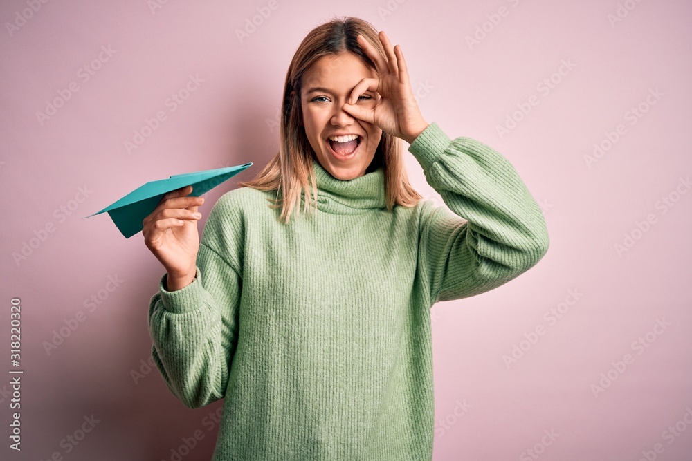 Young beautiful woman holding paper airplane standing over isolated yellow background with happy face smiling doing ok sign with hand on eye looking through fingers Young beautiful woman holding paper airplane standing over isolated yellow background with happy face smiling doing ok sign with hand on eye looking through fingers