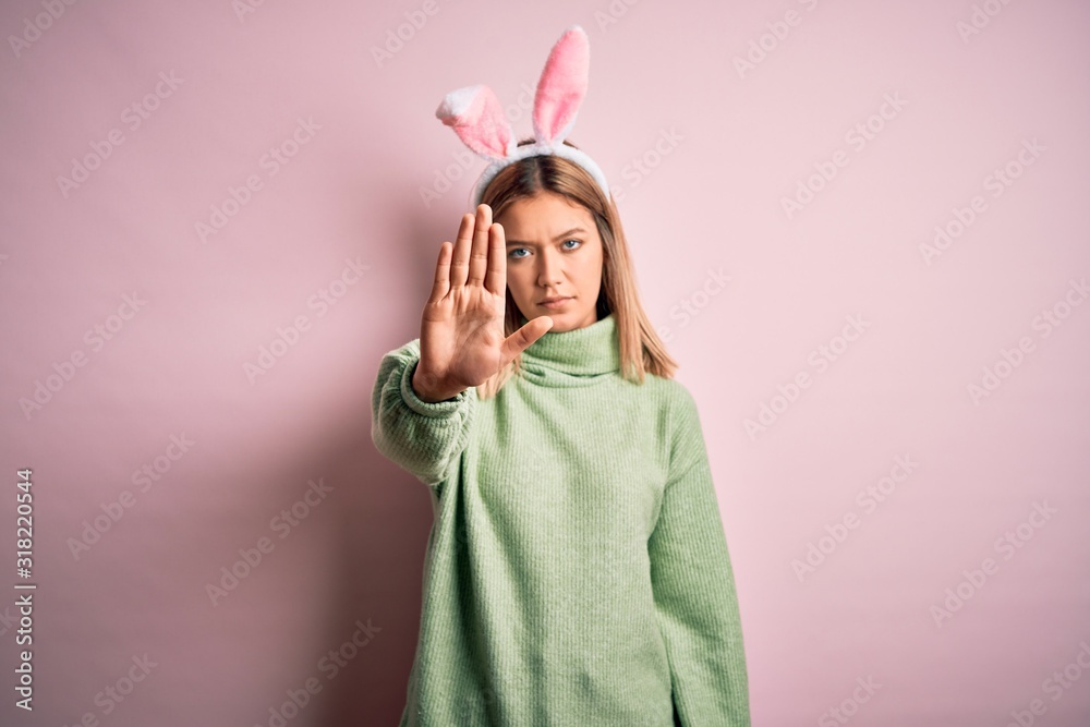 Young beautiful woman wearing easter rabbit ears standing over isolated pink background doing stop sing with palm of the hand. Warning expression with negative and serious gesture on the face.