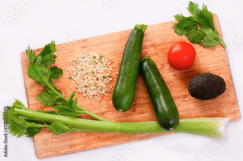 Green vegetables (celery, cucumbers, zucchini, avocado) and a tomato on a wooden chopping board