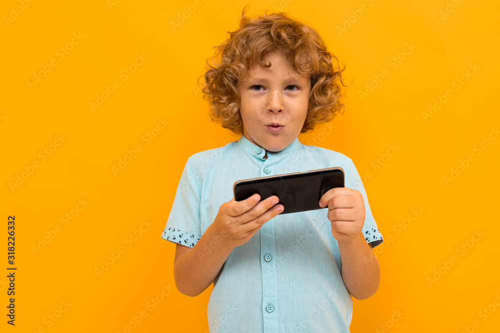 Little boy with curly hair in blue shirt and shorts play phone games isolated on yellow background Little boy with curly hair in blue shirt and shorts play phone games isolated on yellow background