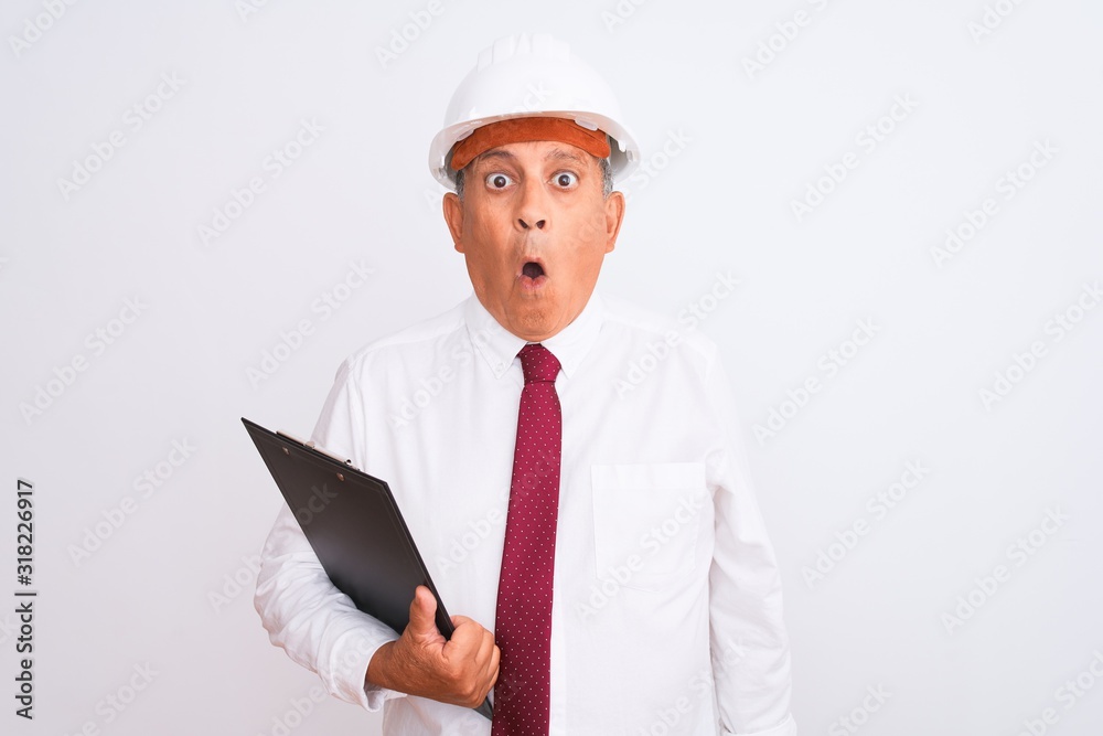 Senior architect man wearing security helmet holding clipboard over isolated white background scared in shock with a surprise face, afraid and excited with fear expression Senior architect man wearing security helmet holding clipboard over isolated white background scared in shock with a surprise face, afraid and excited with fear expression