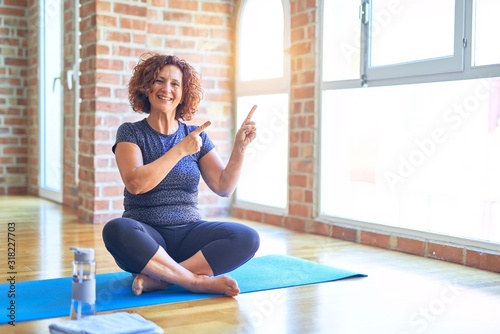 Middle age beautiful sportswoman wearing sportswear sitting on mat practicing yoga at home smiling and looking at the camera pointing with two hands and fingers to the side.