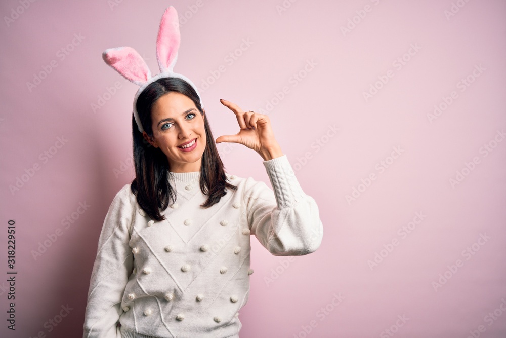 Young caucasian woman wearing cute easter rabbit ears over pink isolated background smiling and confident gesturing with hand doing small size sign with fingers looking and the camera. Measure