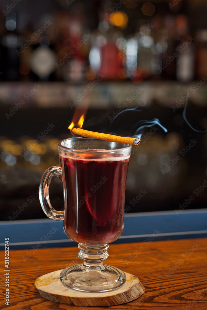 Red cocktail on bar counter. Mulled wine with fruits and cinnamon on the background of bottles with alcohol. Glass with booze on a wooden board. Cinnamon burns and smokes. Red cocktail on bar counter. Mulled wine with fruits and cinnamon on the background of bottles with alcohol. Glass with booze on a wooden board. Cinnamon burns and smokes.