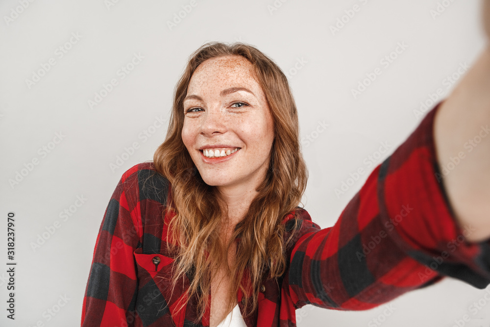 Freestyle. Young woman with freckles standing isolated on grey taking selfie on smartphone posing to camera joyful Freestyle. Young woman with freckles standing isolated on grey taking selfie on smartphone posing to camera joyful