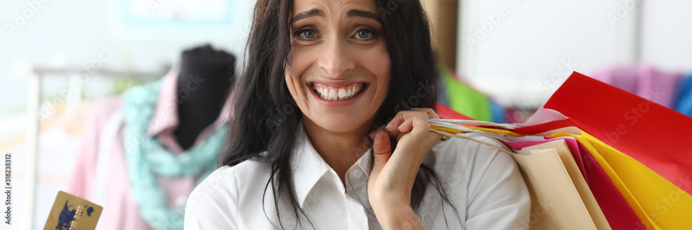 Portrait of smart female standing in fashionable clothes store and showing golden credit card. Beautiful woman holding multicolored packets. Shopping sale concept Portrait of smart female standing in fashionable clothes store and showing golden credit card. Beautiful woman holding multicolored packets. Shopping sale concept