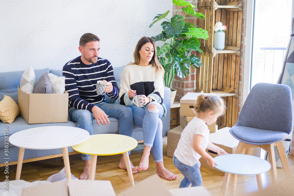 Beautiful family, parents sitting on the sofa drinking coffee looking his kid playing at new home around cardboard boxes Beautiful family, parents sitting on the sofa drinking coffee looking his kid playing at new home around cardboard boxes