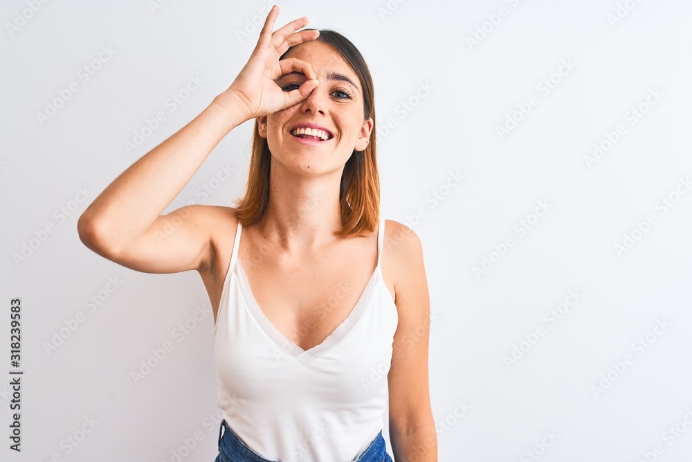 Beautiful redhead woman wearing casual white t-shirt over isolated background doing ok gesture with hand smiling, eye looking through fingers with happy face. Beautiful redhead woman wearing casual white t-shirt over isolated background doing ok gesture with hand smiling, eye looking through fingers with happy face.