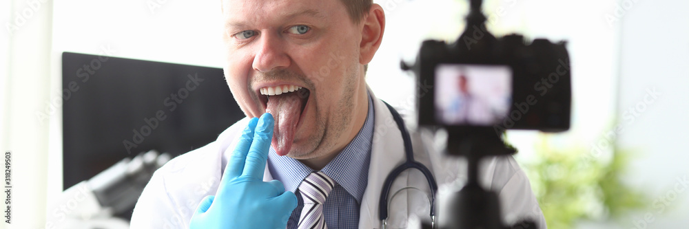 Portrait of joyful practitioner sitting in modern hospital workplace in white uniform with stethoscope and grimacing on high-tech camera. Healthcare and clinic concept Portrait of joyful practitioner sitting in modern hospital workplace in white uniform with stethoscope and grimacing on high-tech camera. Healthcare and clinic concept