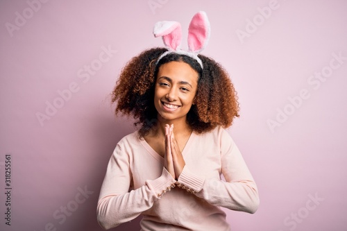 Young african american woman with afro hair wearing bunny ears over pink background praying with hands together asking for forgiveness smiling confident.