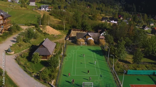 Aerial flight over the football stadium at sunset. Amateur men soccer players playing football outdoors. Beautiful aerial view over the stadium in mountains, near the lake. Soccer field sunset view.