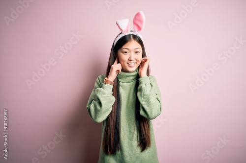 Young asian woman wearing cute easter bunny ears over pink background covering ears with fingers with annoyed expression for the noise of loud music. Deaf concept.