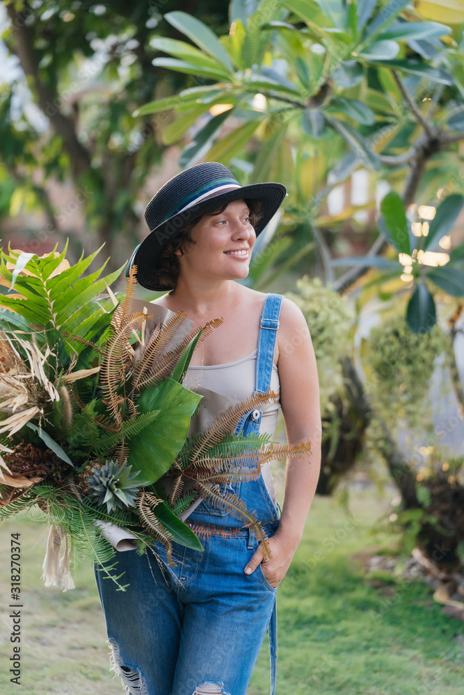 Φωτογραφία Attractive young woman in a hat and denim overalls with a bouquet of tropical flowers posing