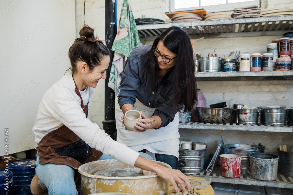 An art teacher giving instruction, help and tips to a student learning to throw clay on a pottery wheel during a lesson. 