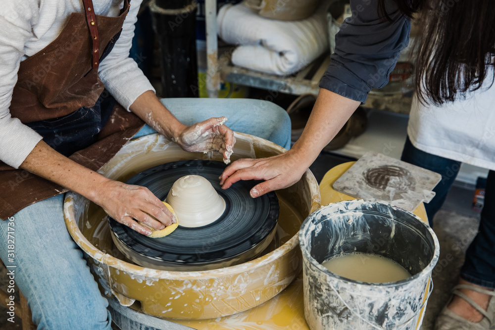A Caucasian woman with a messy bun leaning over a pottery wheel trying to center her clay with a slip bucket nearby.