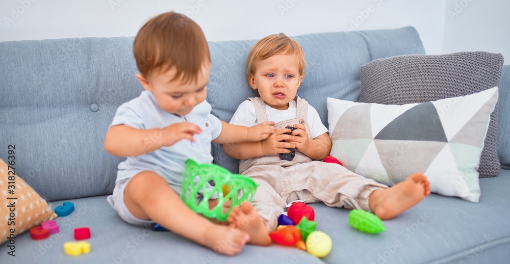 Beautiful toddlers, one of them crying, sitting on the sofa playing with toys at home Beautiful toddlers, one of them crying, sitting on the sofa playing with toys at home