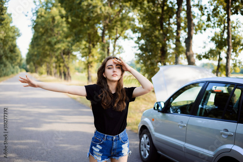 Bewildered caucasian girl puts up a hand to stop approaching car and ask for a help. Car breakdown on the countryside road