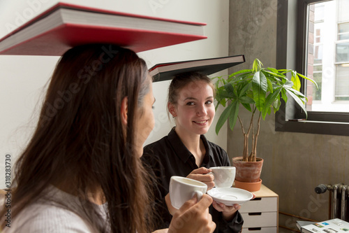 Tableau sur toile Young Women Balancing Books on Top of Heads and Drinking Coffee