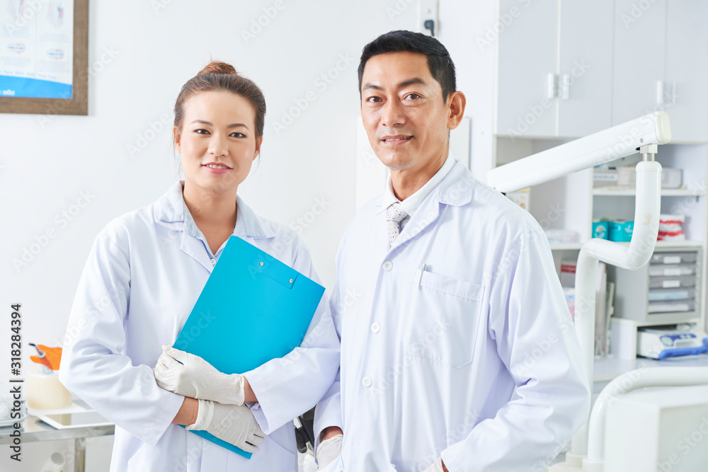 Horizontal medium potrait of dental surgeon and dental assistant wearing white coats looking at camera smiling Horizontal medium potrait of dental surgeon and dental assistant wearing white coats looking at camera smiling
