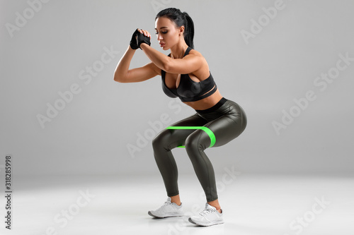 Fitness woman doing squats with resistance band on the gray background. Sporty girl squatting © nikolas_jkd