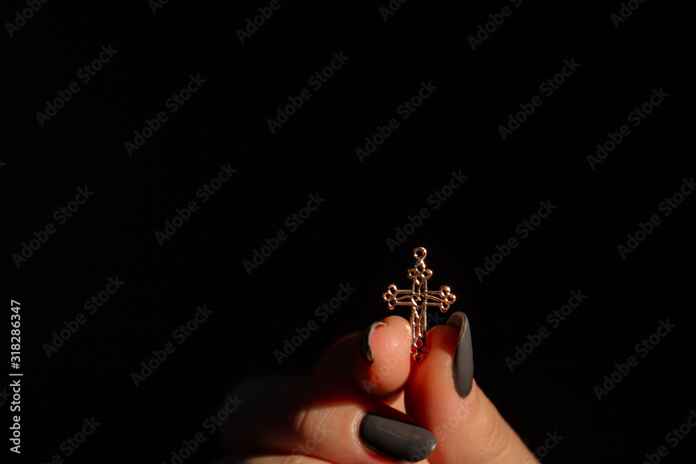 Obraz premium Female's hand is holidng smal golden cross in her fingers. Black background.