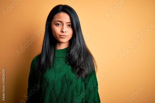 Young beautiful chinese woman wearing green sweater over isolated yellow background with serious expression on face. Simple and natural looking at the camera. © Krakenimages.com