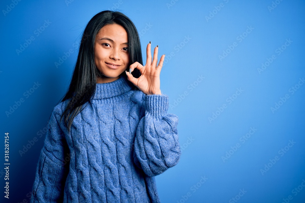 Young beautiful chinese woman wearing casual sweater over isolated blue background smiling positive doing ok sign with hand and fingers. Successful expression.