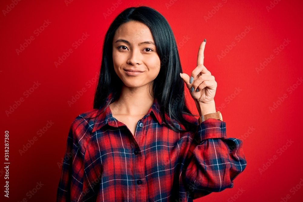 Young beautiful chinese woman wearing casual shirt over isolated red background showing and pointing up with finger number one while smiling confident and happy. Young beautiful chinese woman wearing casual shirt over isolated red background showing and pointing up with finger number one while smiling confident and happy.