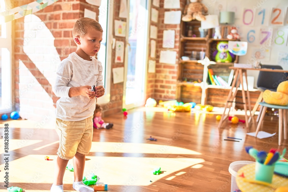 Beautiful blonde toddler playing with small cars around lots of toys at kindergarte