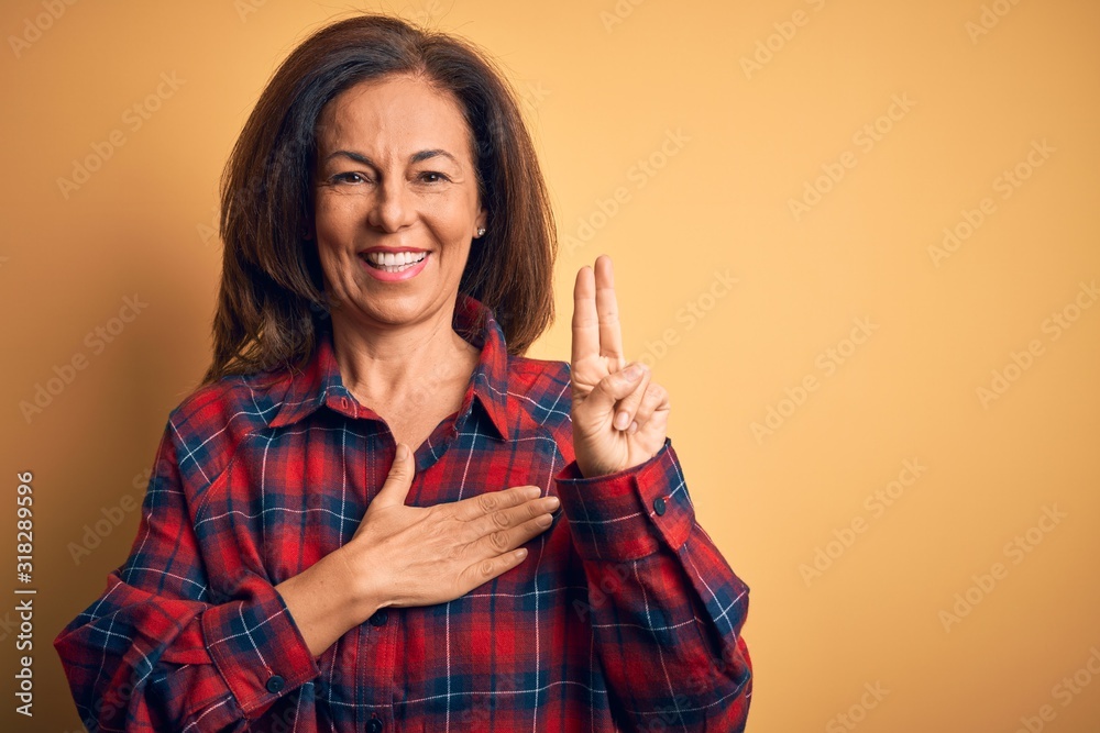 Middle age beautiful woman wearing casual shirt standing over isolated yellow background smiling swearing with hand on chest and fingers up, making a loyalty promise oath Middle age beautiful woman wearing casual shirt standing over isolated yellow background smiling swearing with hand on chest and fingers up, making a loyalty promise oath
