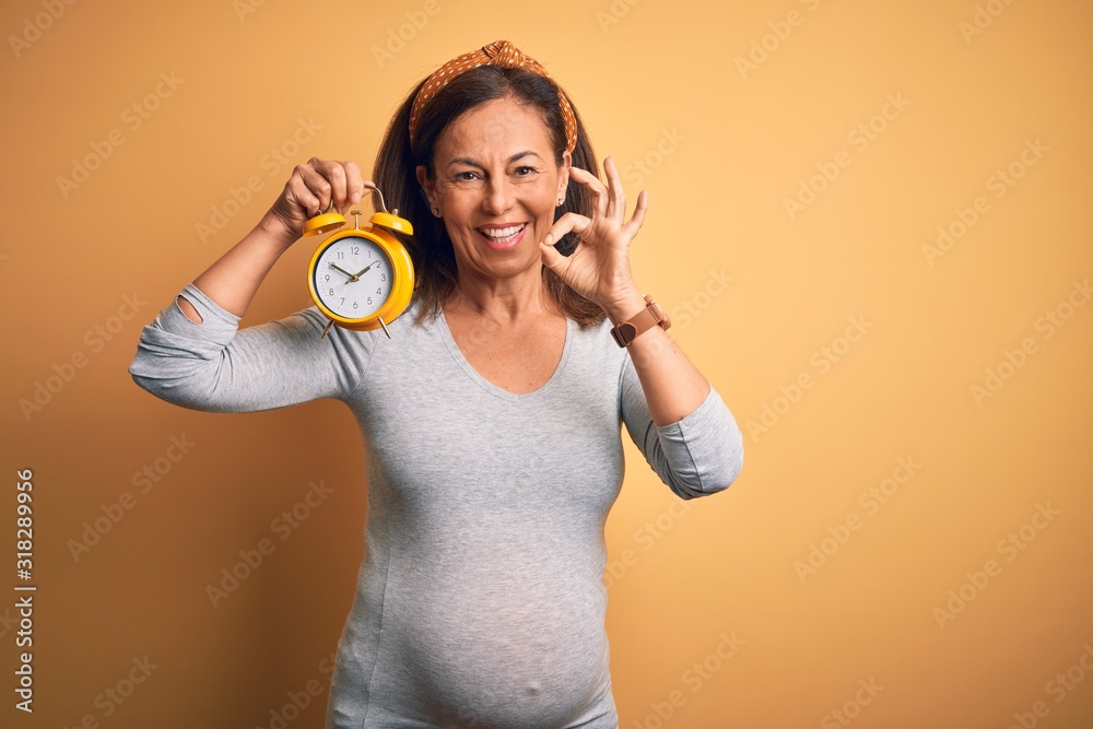 Obraz premium Middle age pregnant woman expecting baby at aged pregnancy holding alarm clock doing ok sign with fingers, excellent symbol