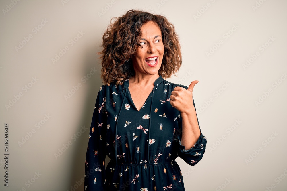 Middle age beautiful woman wearing casual dress standing over isolated white background smiling with happy face looking and pointing to the side with thumb up. Middle age beautiful woman wearing casual dress standing over isolated white background smiling with happy face looking and pointing to the side with thumb up.