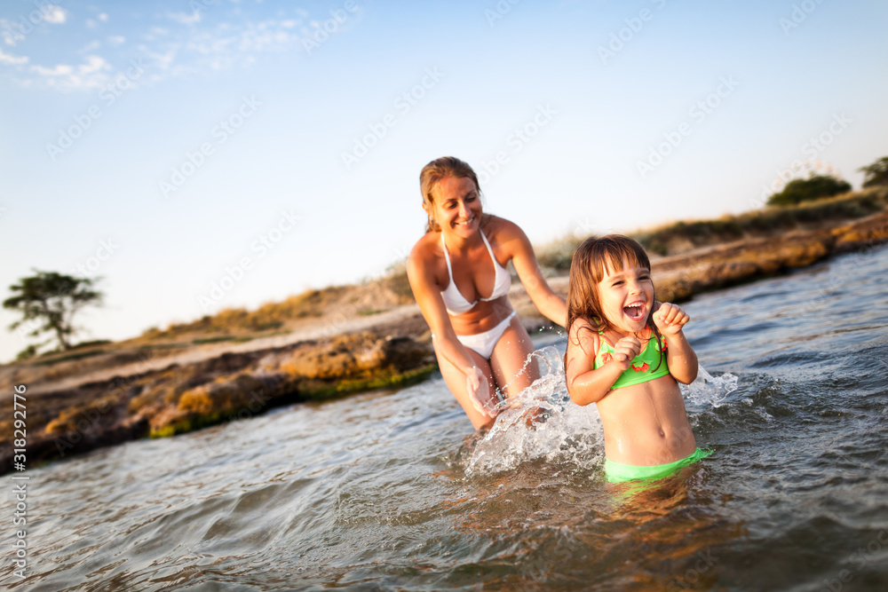 Young blond woman in white bikini and small happy girl having fun in sea water with rocky beach at background on clear summer day. Travelling, vacations, relaxation, family weekend concept
