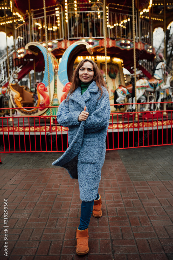 Young stylish woman in a fashionable faux fur coat in a trendy blue collor 2020 near a carousel in the city.