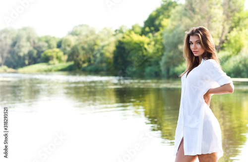 Young sexy brunette woman in white dress standing near river and enjoying sunshine on summer day