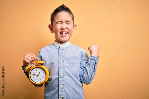 Young little boy kid holding classic bell alarm clock over isolated yellow background screaming proud and celebrating victory and success very excited, cheering emotion