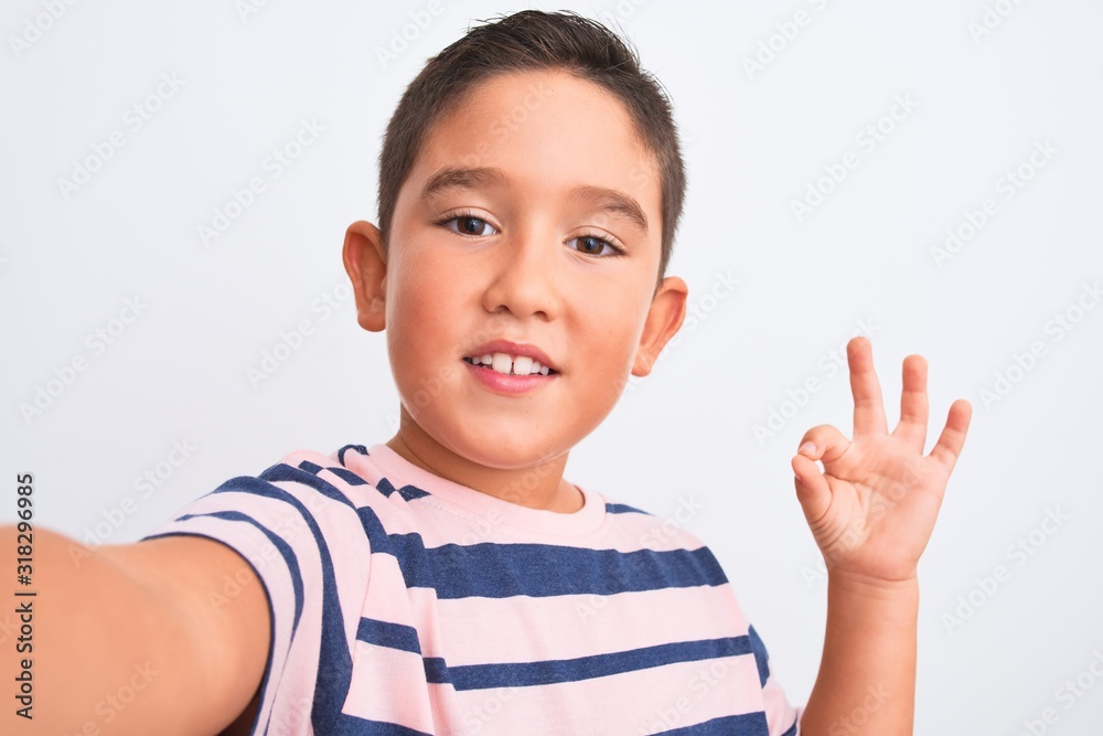 Beautiful kid boy wearing casual striped t-shirt make selfie over isolated white background doing ok sign with fingers, excellent symbol Beautiful kid boy wearing casual striped t-shirt make selfie over isolated white background doing ok sign with fingers, excellent symbol