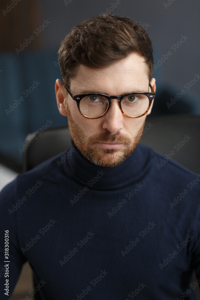 Young businessman in office. Business portrait of handsome bearded man wearing eyeglasses sitting at workplace. Confident businessman became successful. Business concept. Young businessman in office. Business portrait of handsome bearded man wearing eyeglasses sitting at workplace. Confident businessman became successful. Business concept.