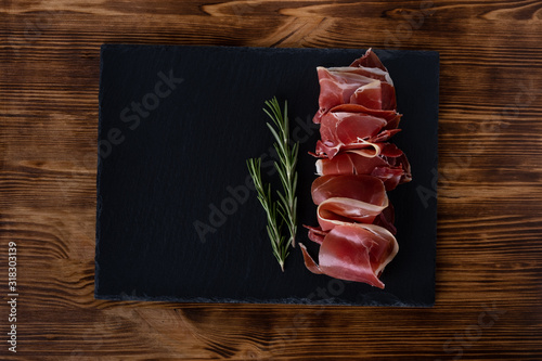 Prosciutto crudo or jamon with rosemary on black board on wooden background, top view. Italian antipasto. Traditional Spanish Jamon Serrano ham, Prosciutto Crudo, Parma ham