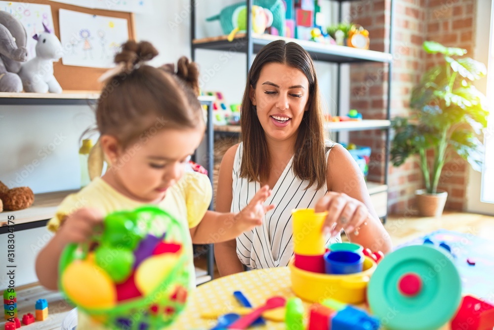 Fototapeta premium Young beautiful teacher and toddler playing with dishes, cutlery and cups toy on the table at kindergarten