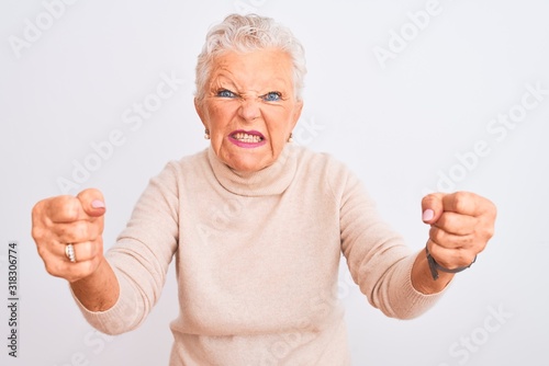 Senior grey-haired woman wearing turtleneck sweater standing over isolated white background angry and mad raising fists frustrated and furious while shouting with anger. Rage and aggressive concept.