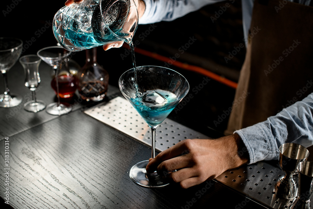 Bartender pouring a blue color alcoholic cocktail from the measuring cup with strainer to a martini glass