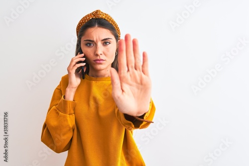 Young beautiful woman talking on the smartphone standing over isolated white background with open hand doing stop sign with serious and confident expression, defense gesture
