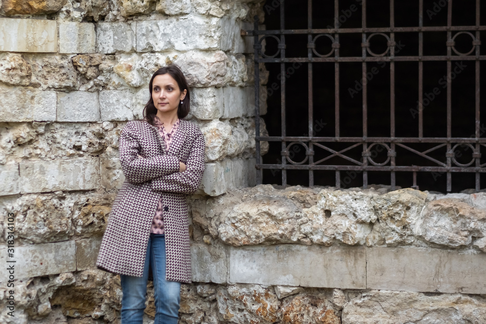 Young brunette woman standing near stone wall with metal fence. Pretty model portrait on old building background.