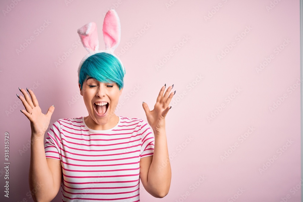 Young woman with fashion blue hair wearing easter rabbit ears over pink background celebrating mad and crazy for success with arms raised and closed eyes screaming excited. Winner concept Young woman with fashion blue hair wearing easter rabbit ears over pink background celebrating mad and crazy for success with arms raised and closed eyes screaming excited. Winner concept