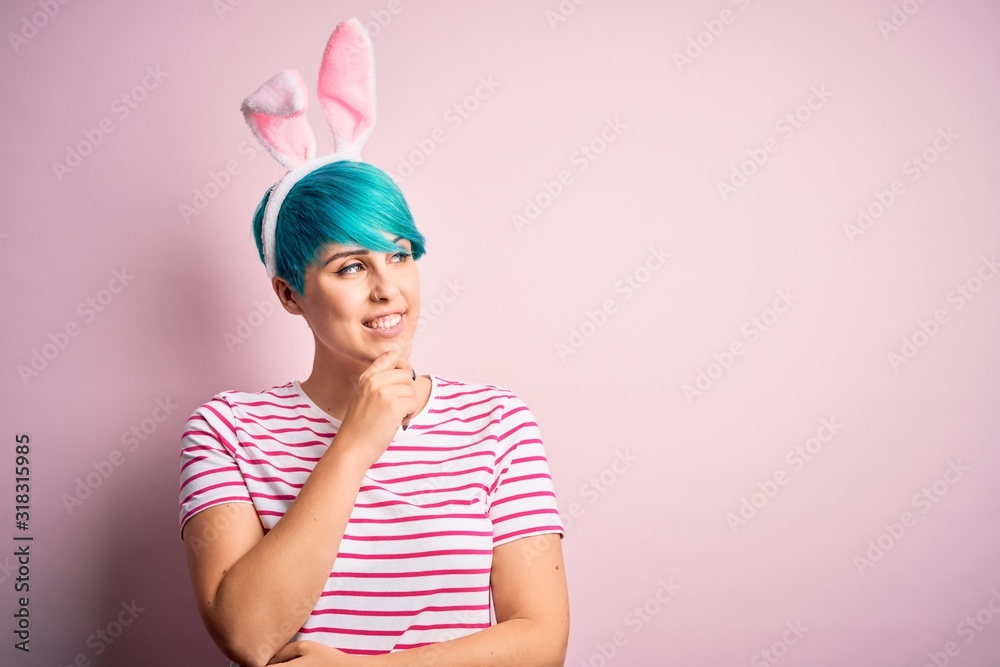 Young woman with fashion blue hair wearing easter rabbit ears over pink background with hand on chin thinking about question, pensive expression. Smiling with thoughtful face. Doubt concept.