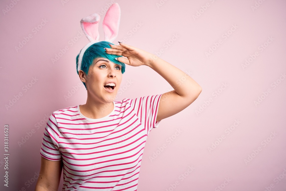 Young woman with fashion blue hair wearing easter rabbit ears over pink background very happy and smiling looking far away with hand over head. Searching concept. Young woman with fashion blue hair wearing easter rabbit ears over pink background very happy and smiling looking far away with hand over head. Searching concept.